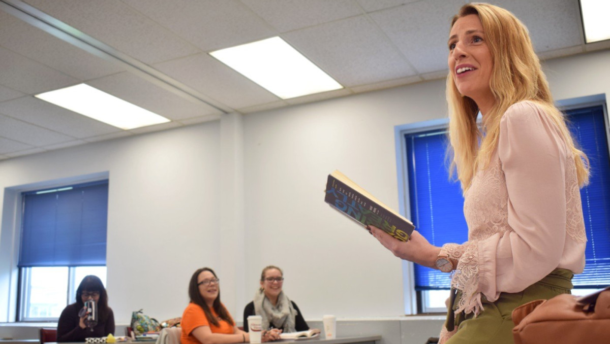 Instructor holding a book while speaking to students seated in a classroom. Identified as informative because the image depicts a teaching interaction that provides instructional context.