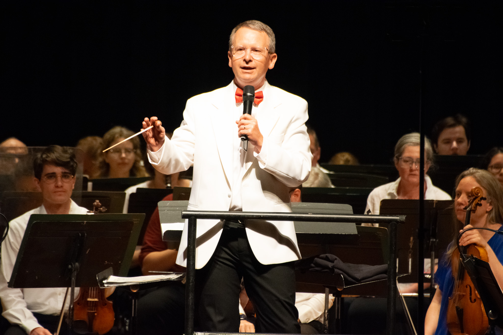 A conductor in a white jacket and red bow tie speaks into a microphone while standing in front of an orchestra during a live concert performance.