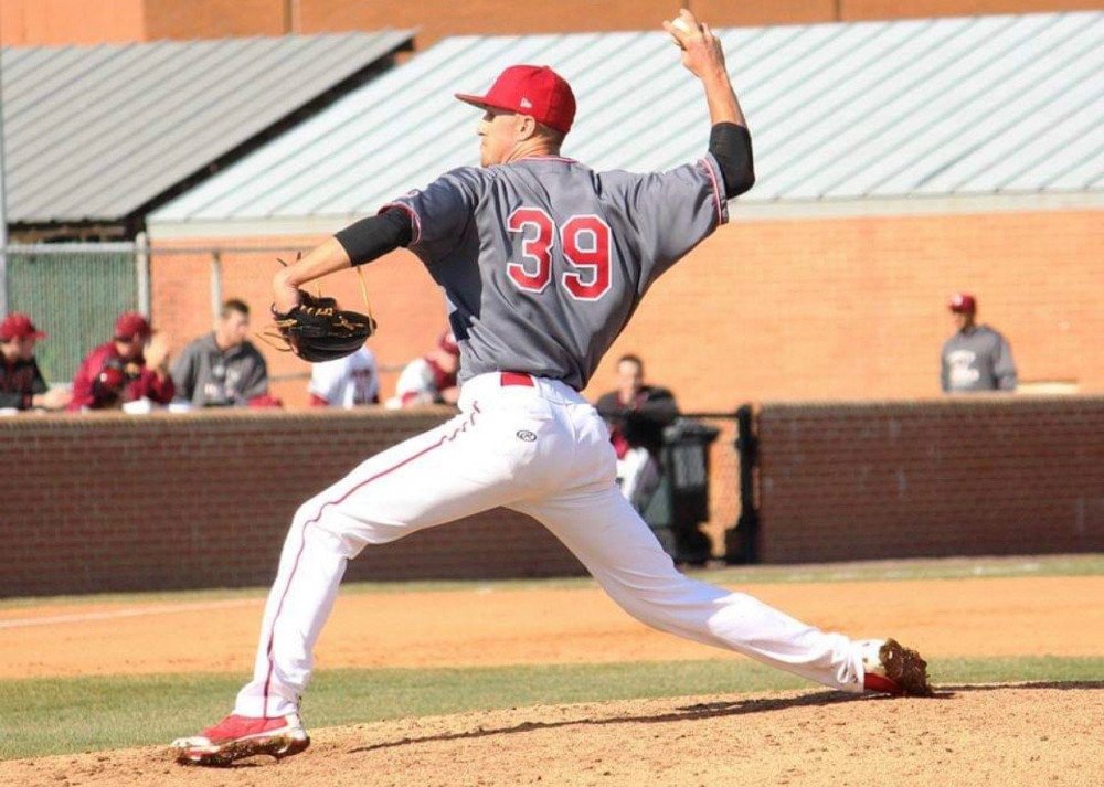 Joe Kuzia throws a baseball to his left while on the pitcher's mound. He's dressed in a gray and red jersey.