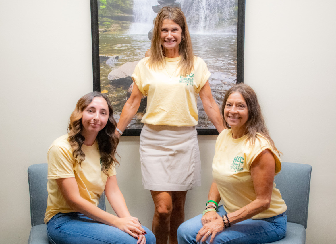 Three women wearing matching yellow “HCCC Generals Welcome Crew” shirts smile for a group photo in a seating area, with one woman standing between two seated women and a framed waterfall photograph behind them.